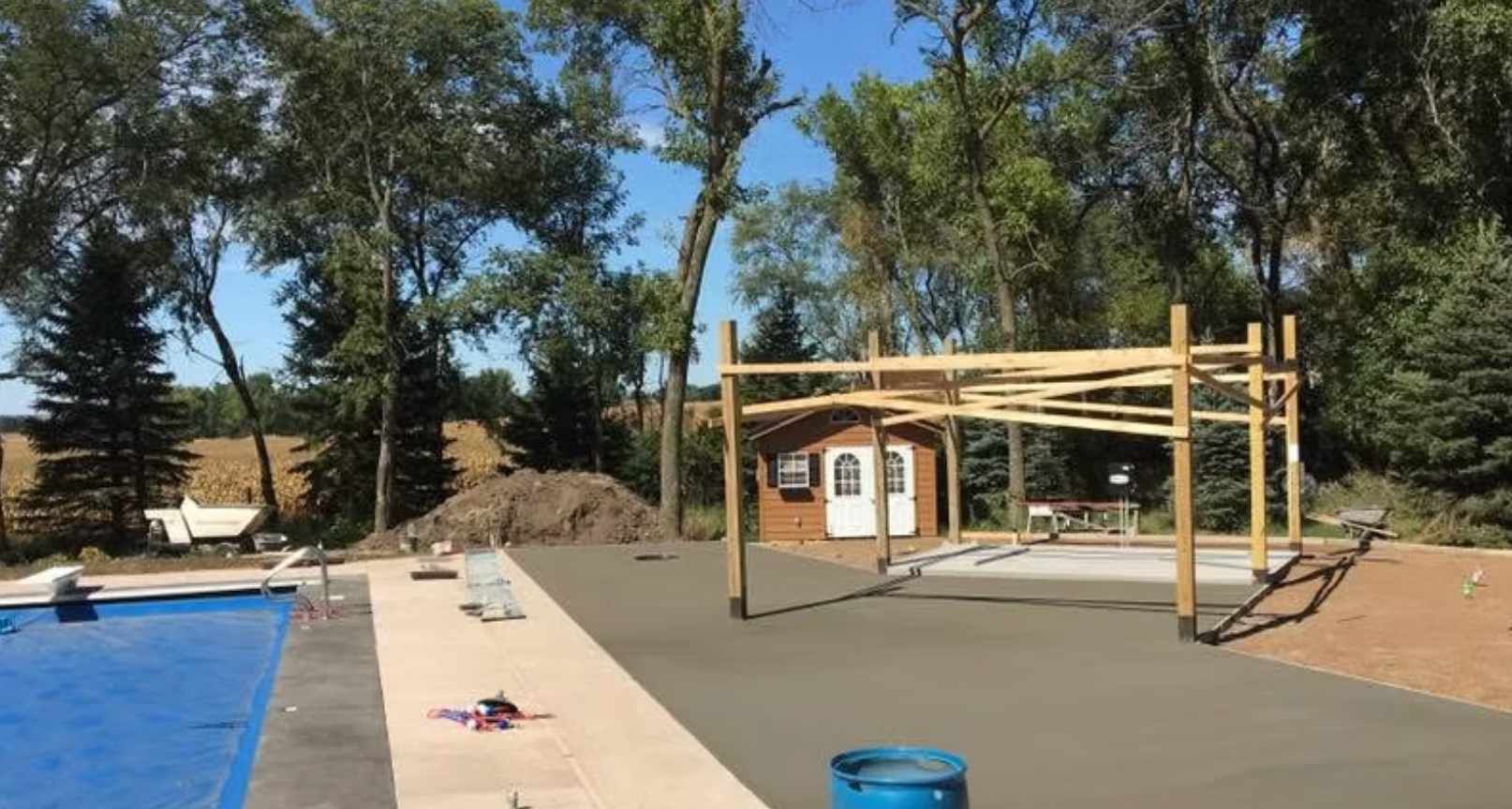 Backyard poolside deck with a wooden pergola, chairs, trees, and a small shed in the background