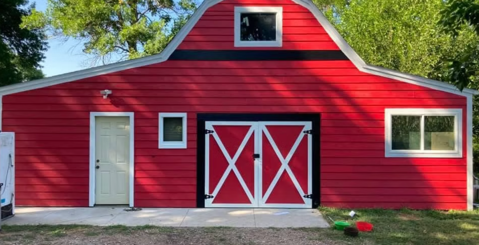 Red barn with white trim, double doors, and a side door in a grassy yard under trees.