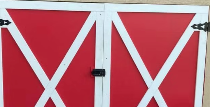 Red barn double doors with white X trim and black hinges, closed on a white wall