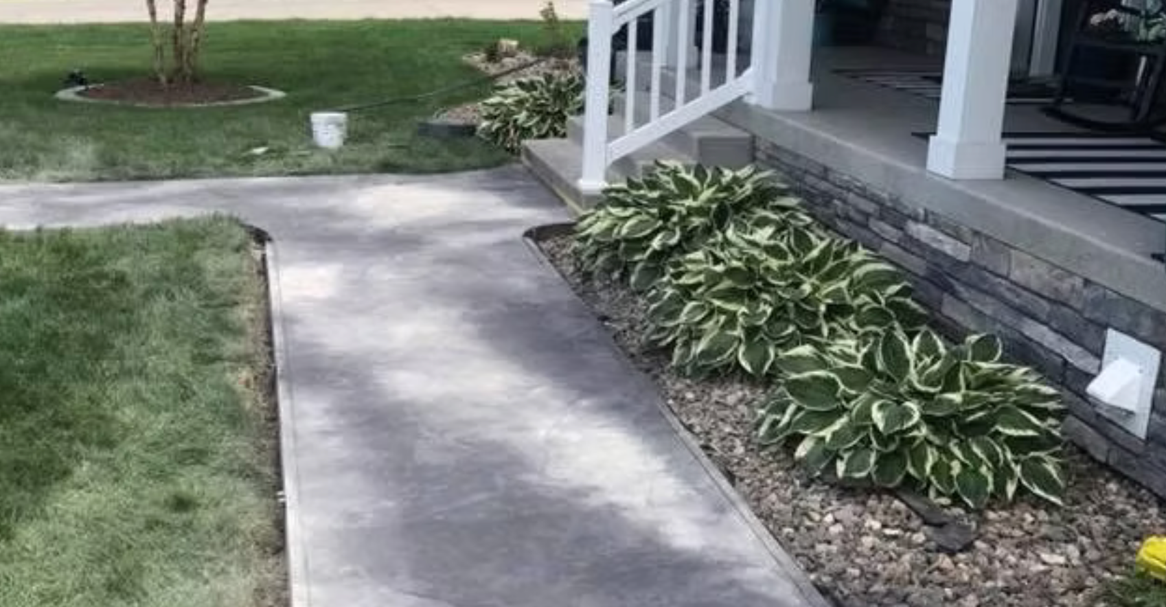 Concrete walkway beside a porch with steps and potted plants, bordered by grass and a hosta bed