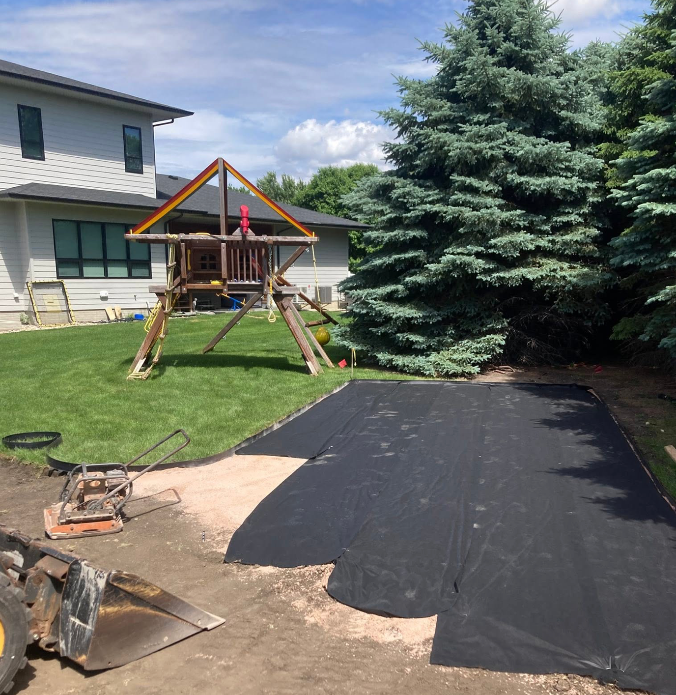 Backyard with a wooden playset, large evergreens, and a black tarp-covered area beside a house.
