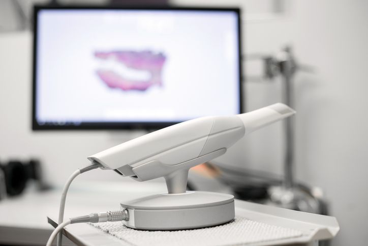 Dental scanner on a table with a blurred monitor displaying a mouth scan in the background