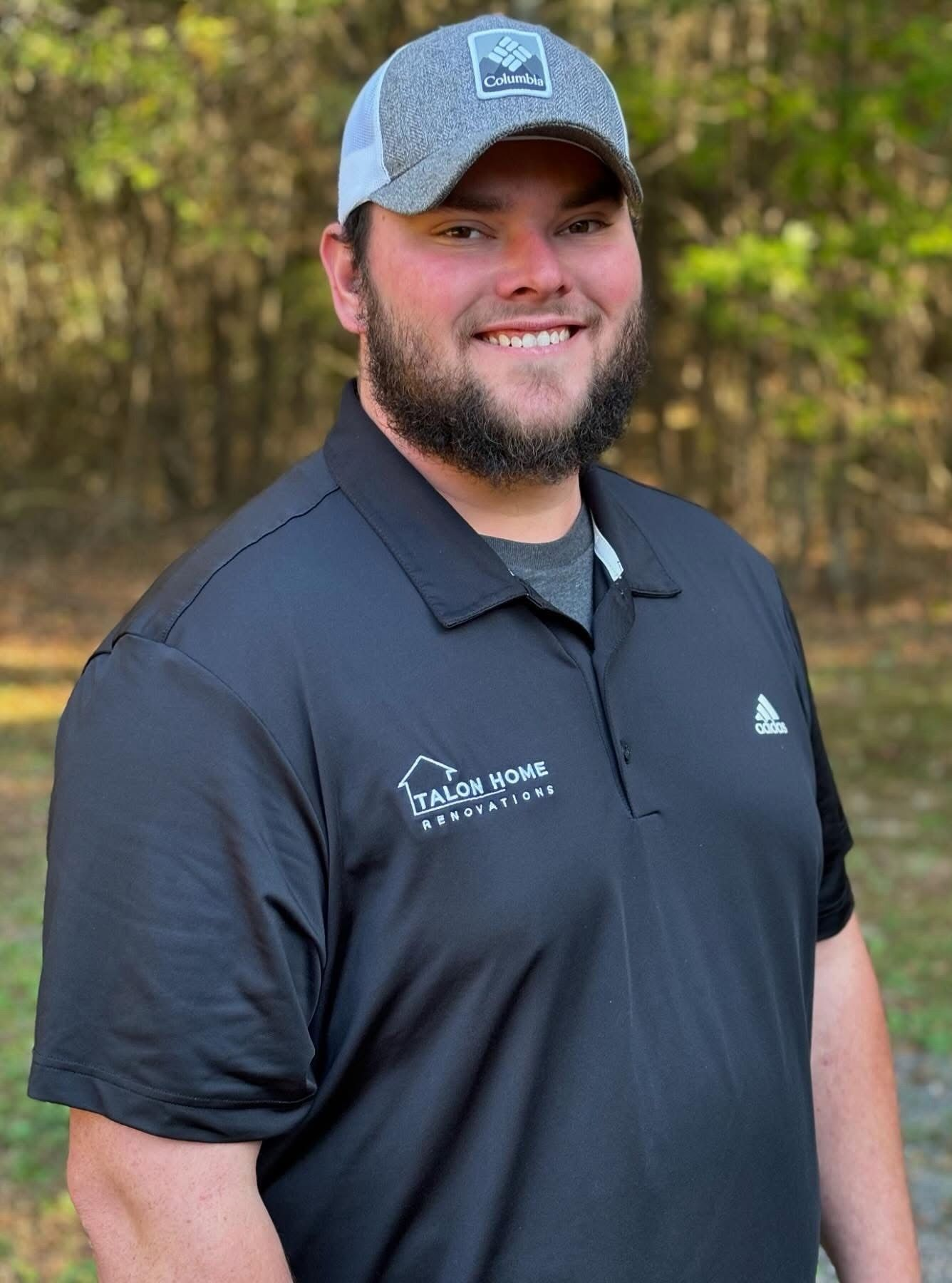 Smiling man in a navy polo and gray cap standing outdoors in front of trees