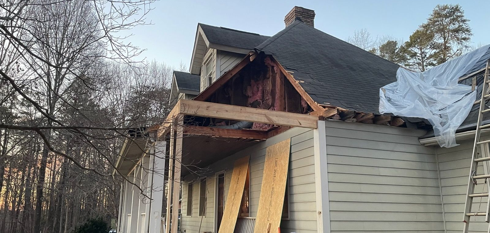 Partially demolished house with exposed roof framing, boarded front, and debris-covered side.