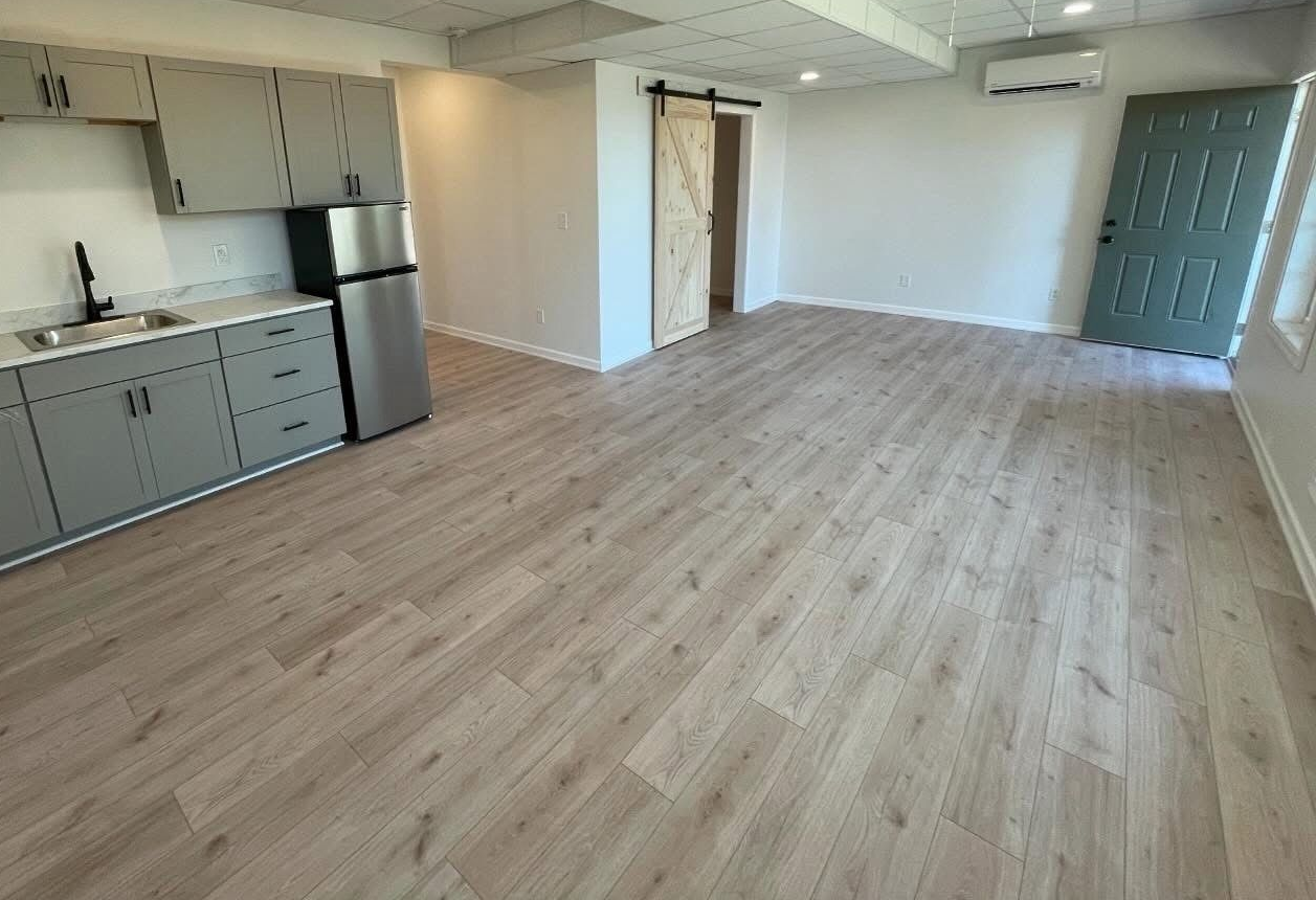 Empty open-plan kitchen and living area with light wood floors, gray cabinets, and a blue door