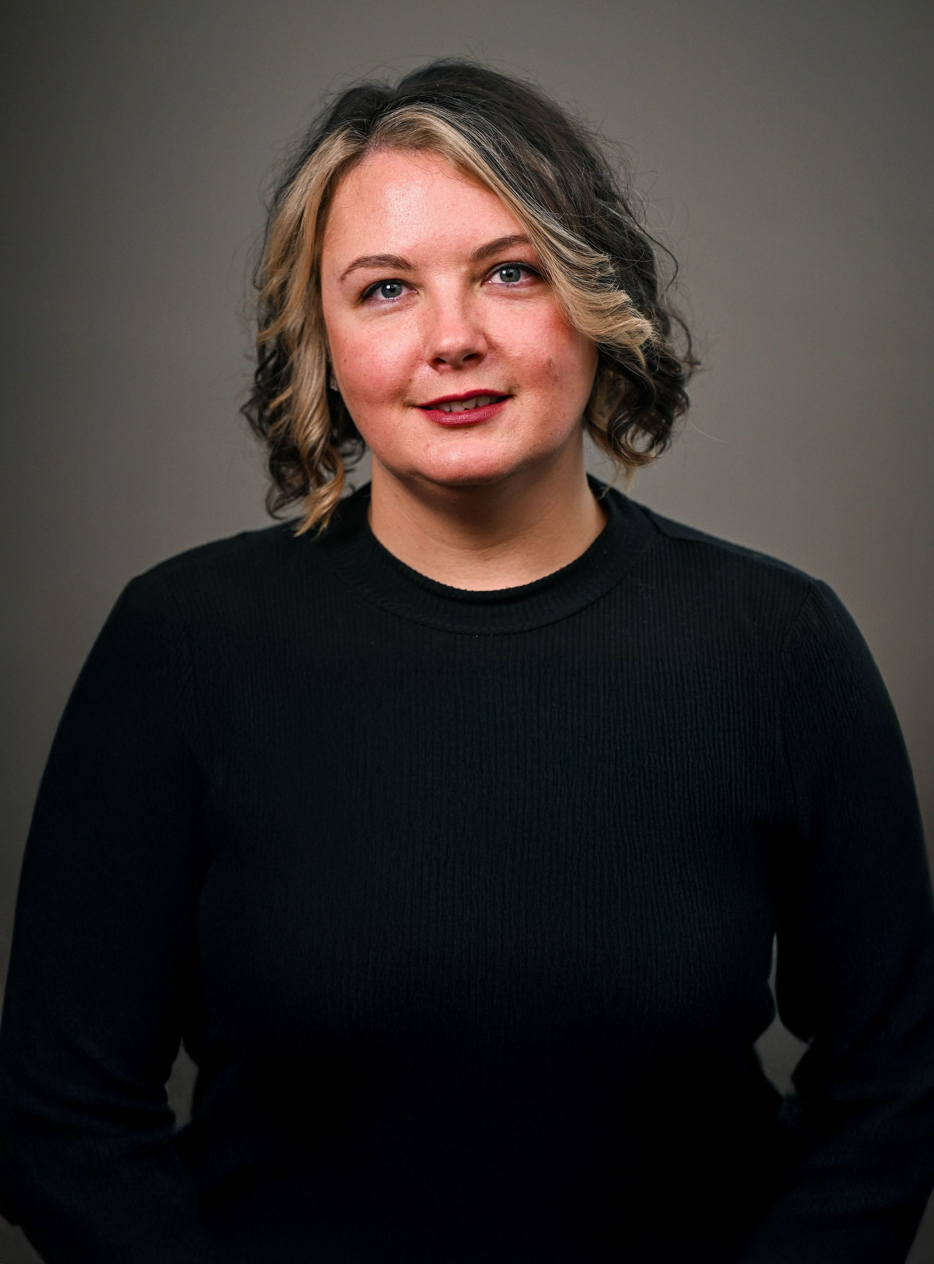 Woman with wavy, highlighted hair smiles, wearing a black sweater against a gray backdrop.