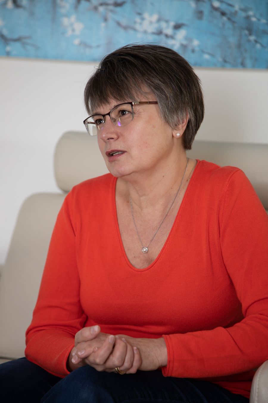 Woman in a red top speaking on a couch, with a blue painting in the background.