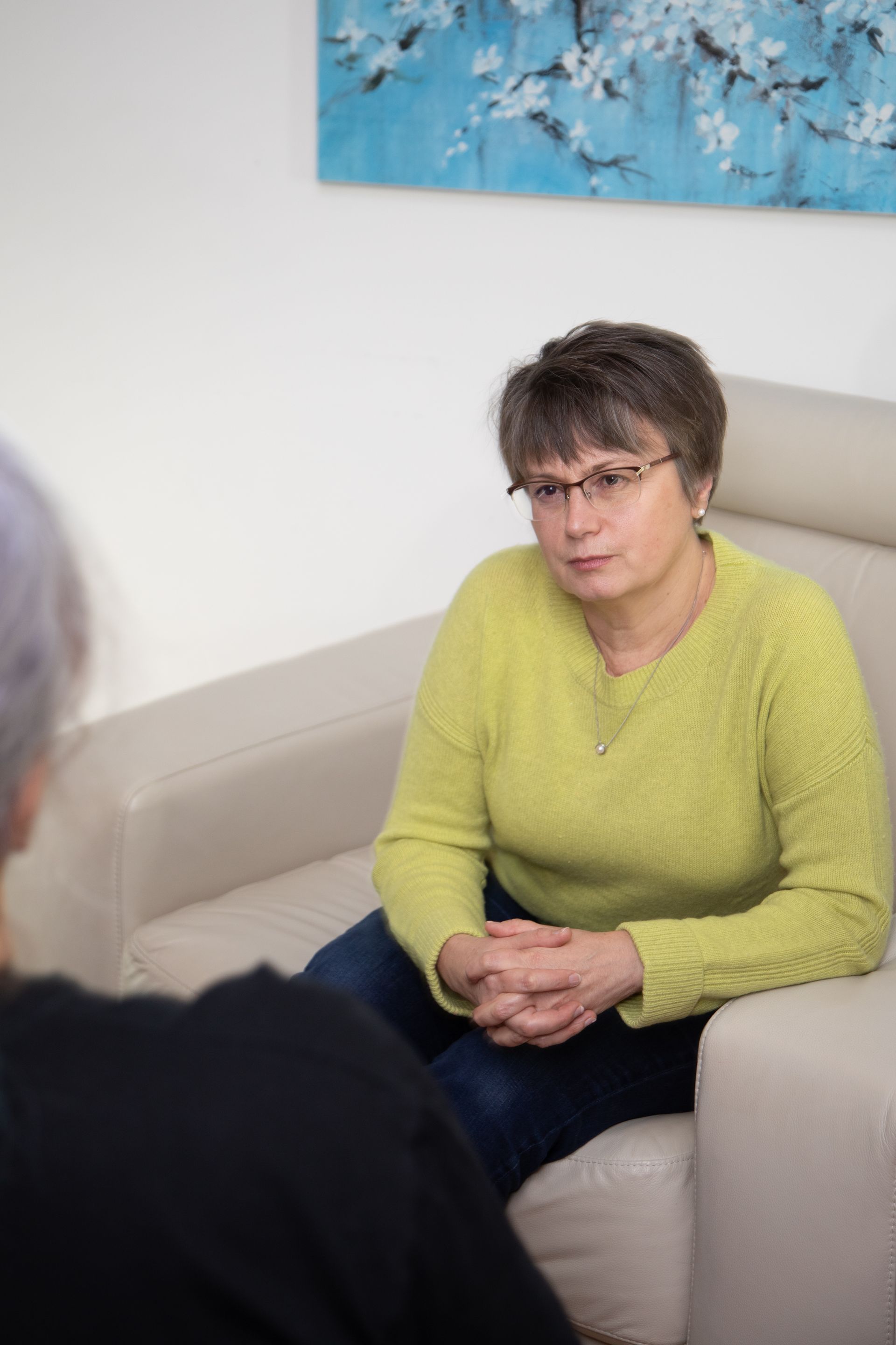 Woman in a lime-green sweater sits on a beige couch, speaking with someone in a counseling setting.
