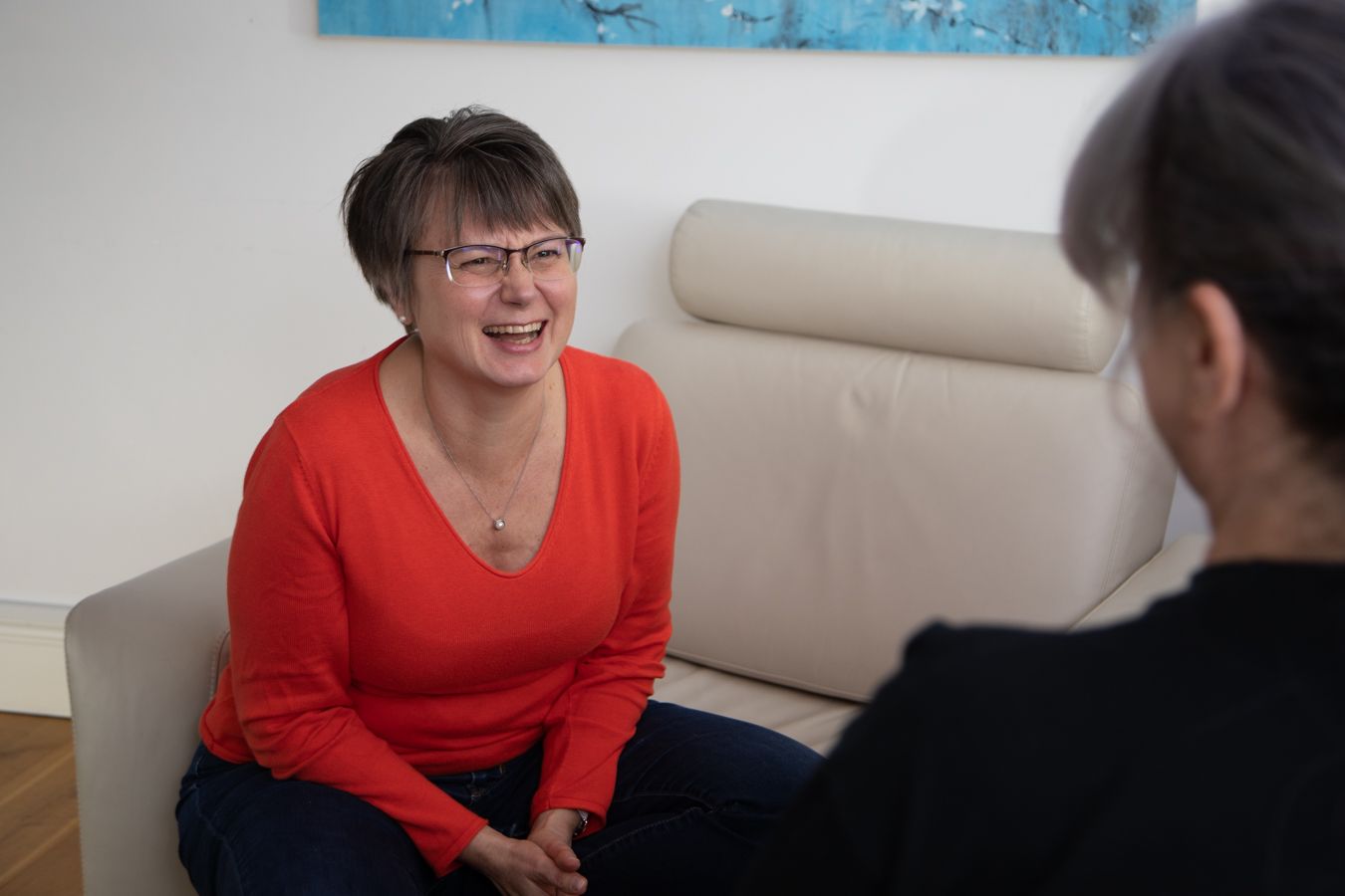 Two people sitting and talking on a beige couch; one smiles in a red top.