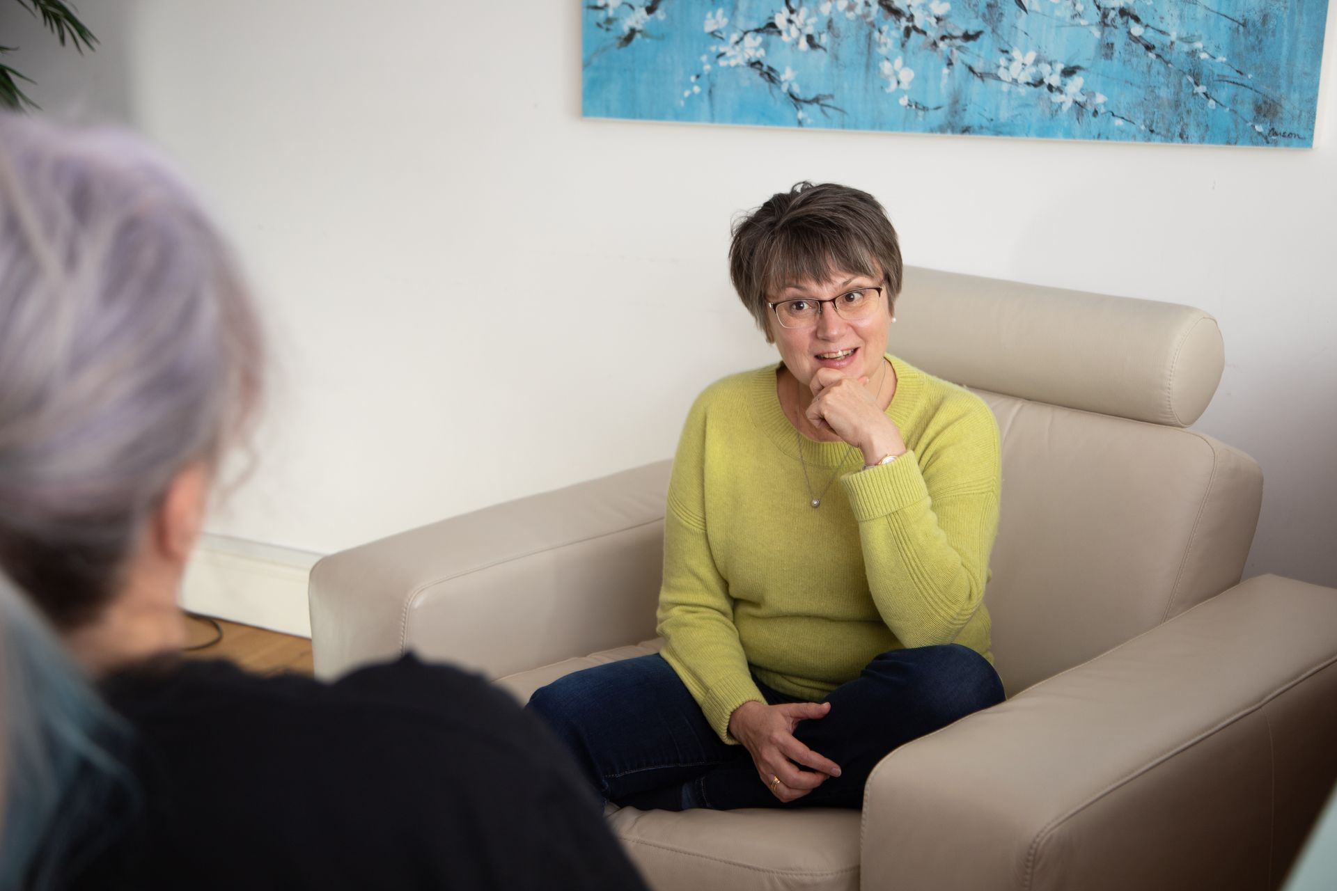 Woman in green sweater seated on beige couch, listening during a conversation in a bright room.