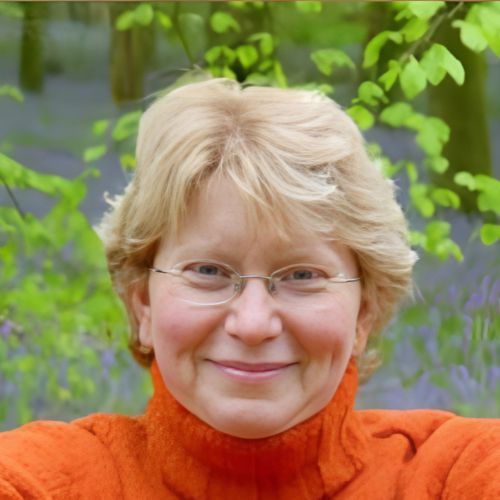 Smiling person with glasses in an orange sweater, outdoors with green leaves and purple flowers behind them