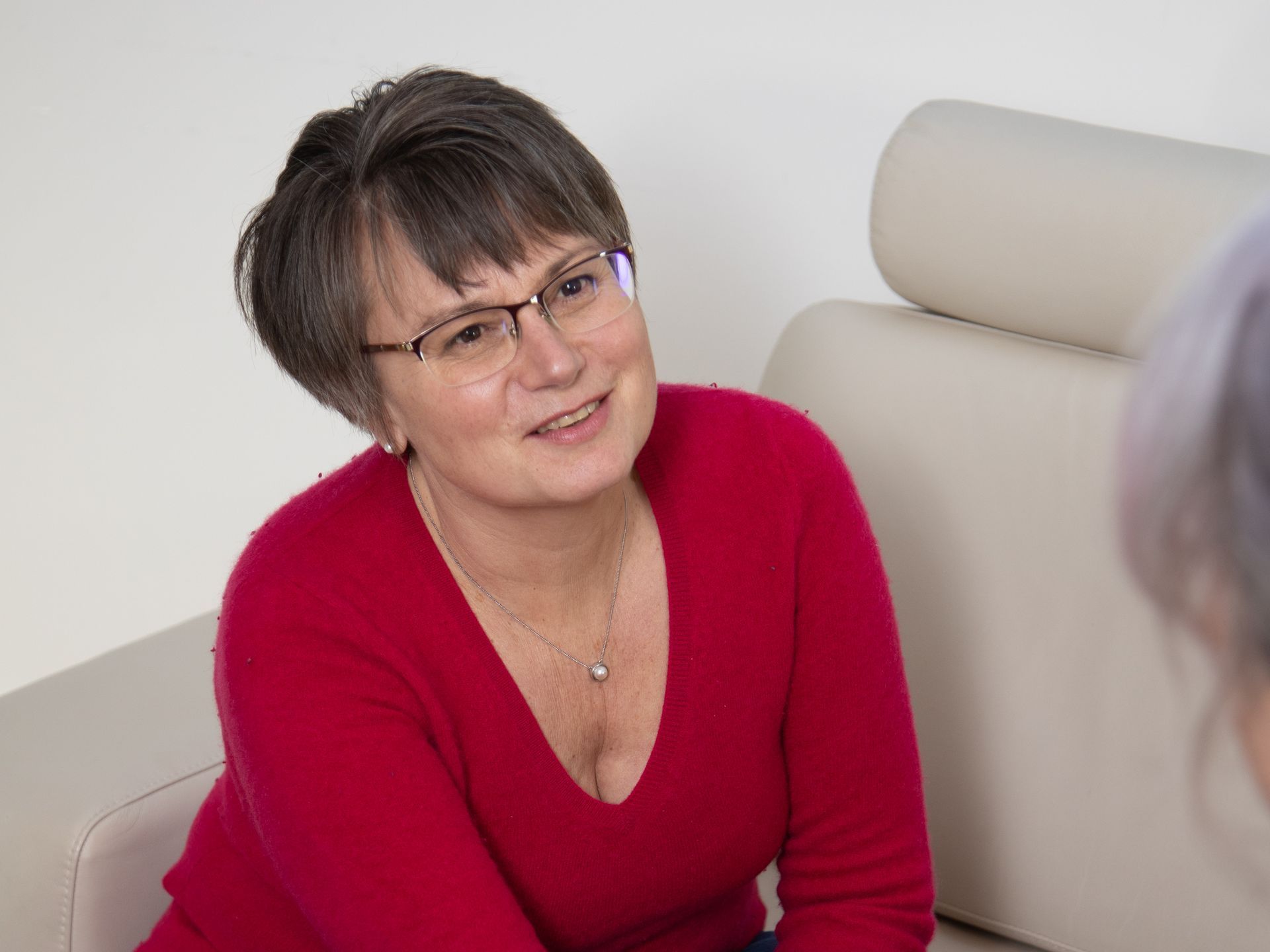 Smiling woman in a red top and glasses talking on a beige sofa