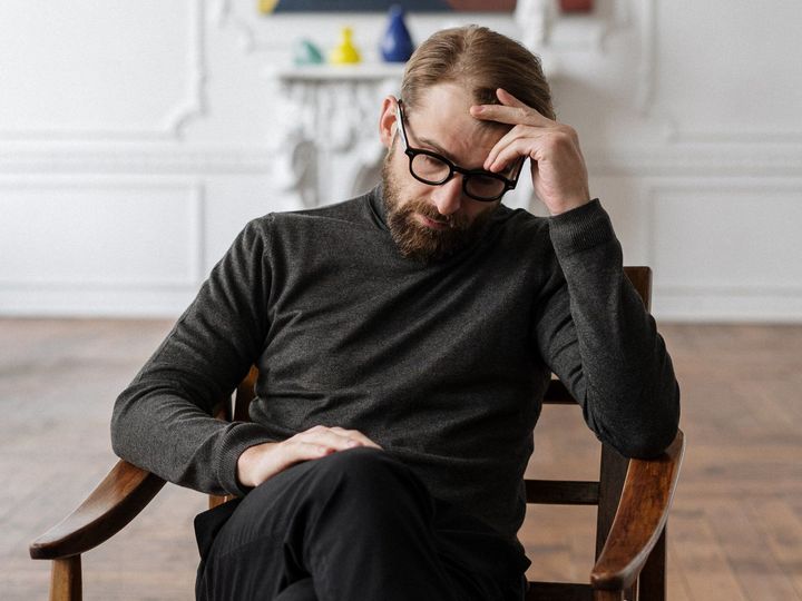 Man in glasses sitting on a wooden chair, resting his head in his hand, looking pensive in a bright room