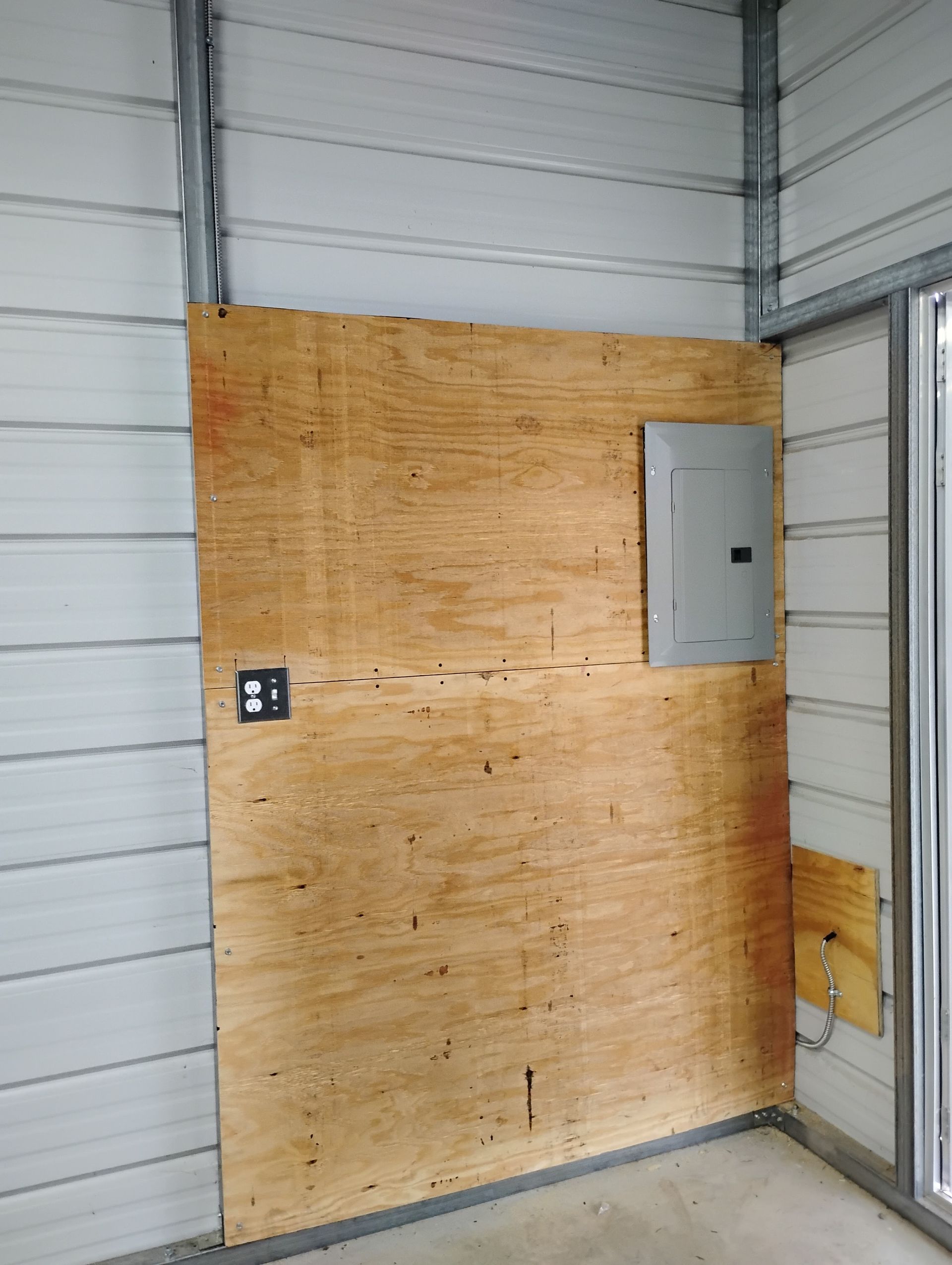 An electrical panel and outlet mounted on a plywood sheet attached to the interior wall of a metal building.
