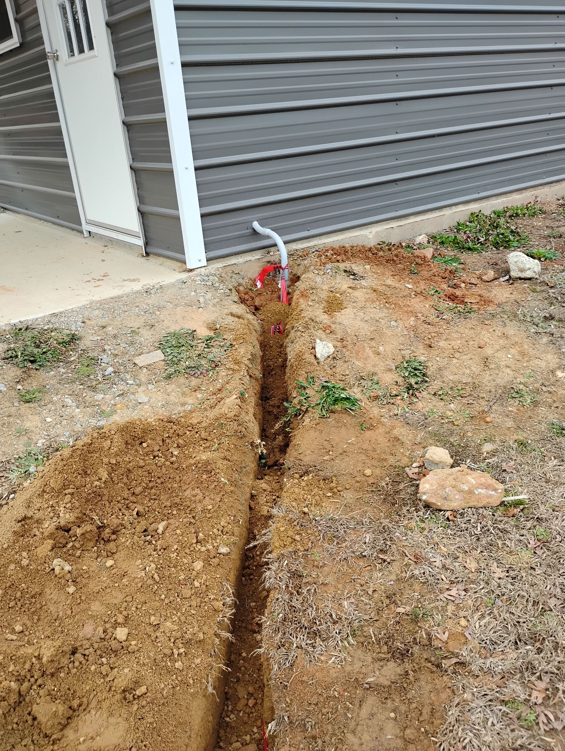 A narrow trench runs through dirt and grass alongside a building with gray metal siding and a white door.
