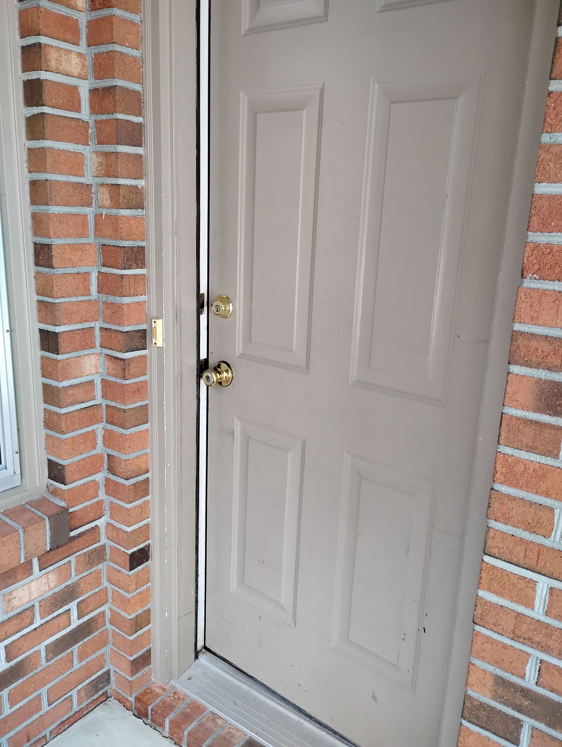 A slightly ajar beige door with gold hardware set into a brick wall.