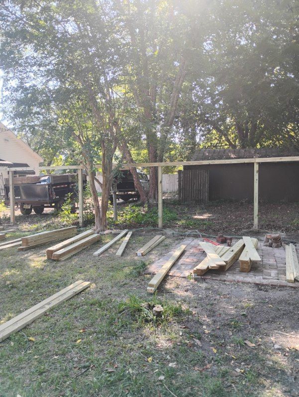 Outdoor yard with scattered lumber, trees, and a small shed or carport in the background