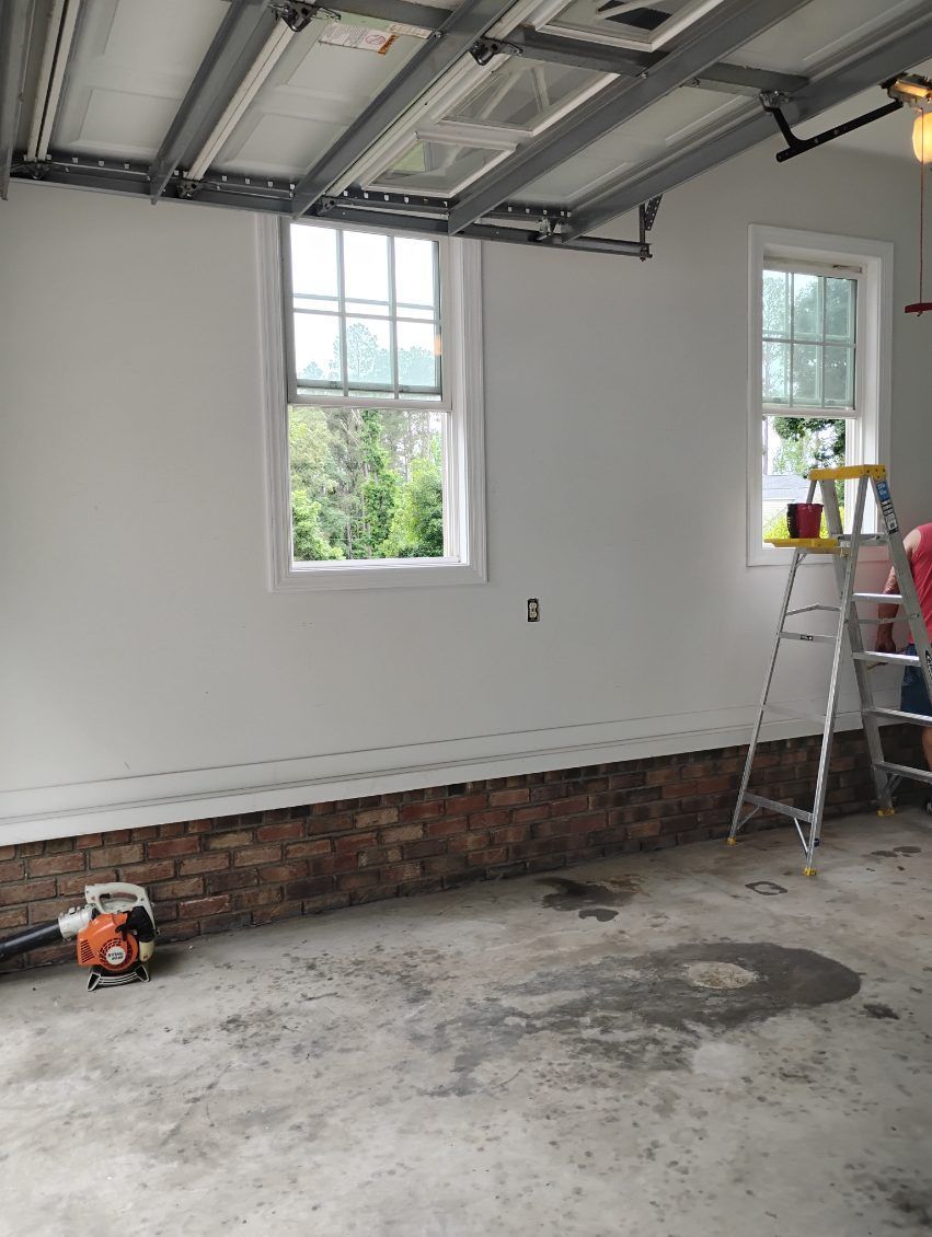 Empty garage under renovation with open garage door, brick wall, windows, ladder, and tools on concrete floor