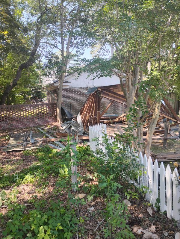 Rustic yard with fallen wooden fence, white picket fence, trees, and a small shed in the background