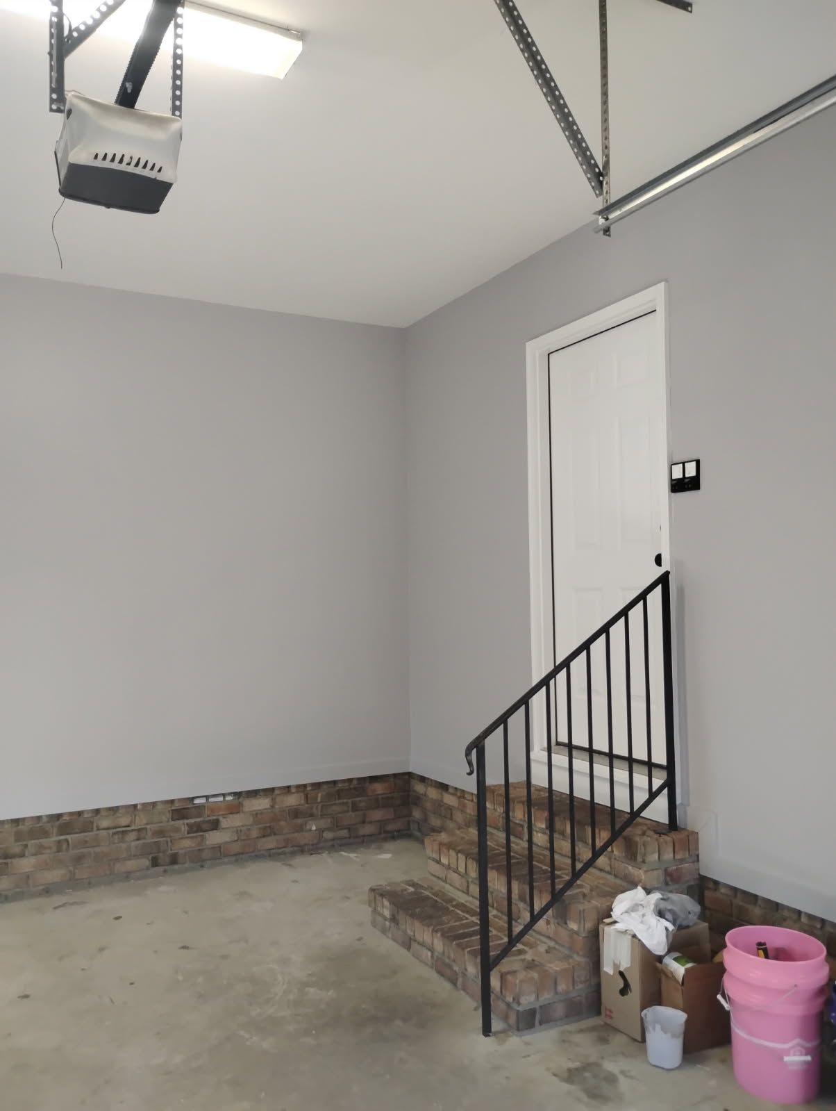 Empty garage with gray walls, concrete floor, brick trim, stairs, and a white side door.