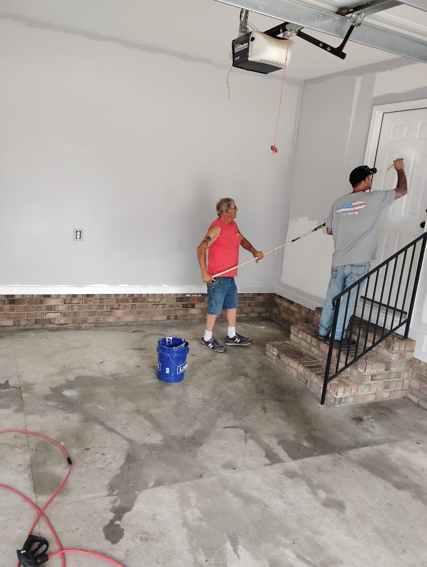 Two people painting a garage interior with a ladder, bucket, and roller.