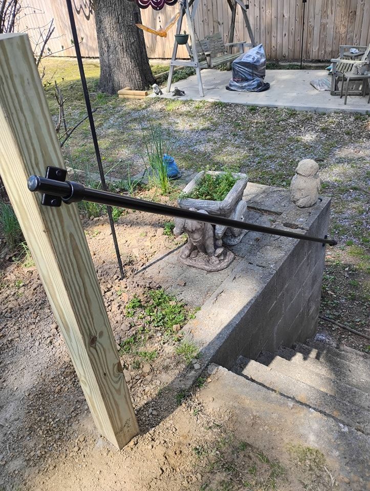 A black metal handrail bracket attached to a vertical wooden post overlooking a concrete staircase in a backyard.