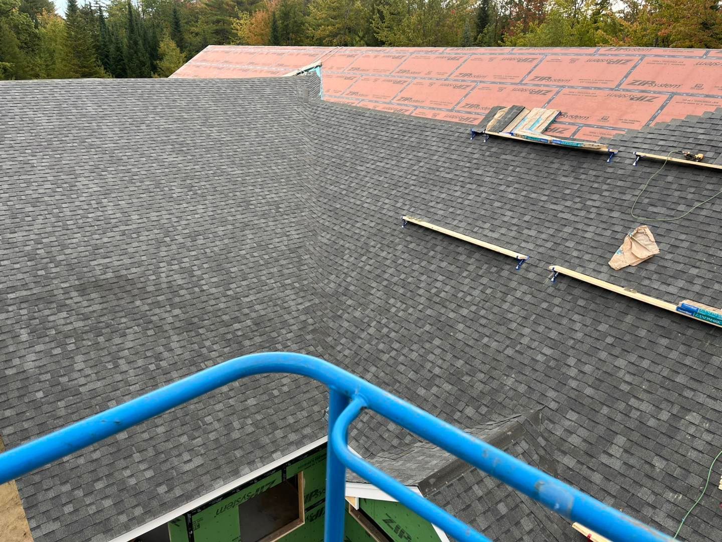 A blue metal safety railing overlooks a roof under construction, featuring sections of dark shingles and red underlayment.