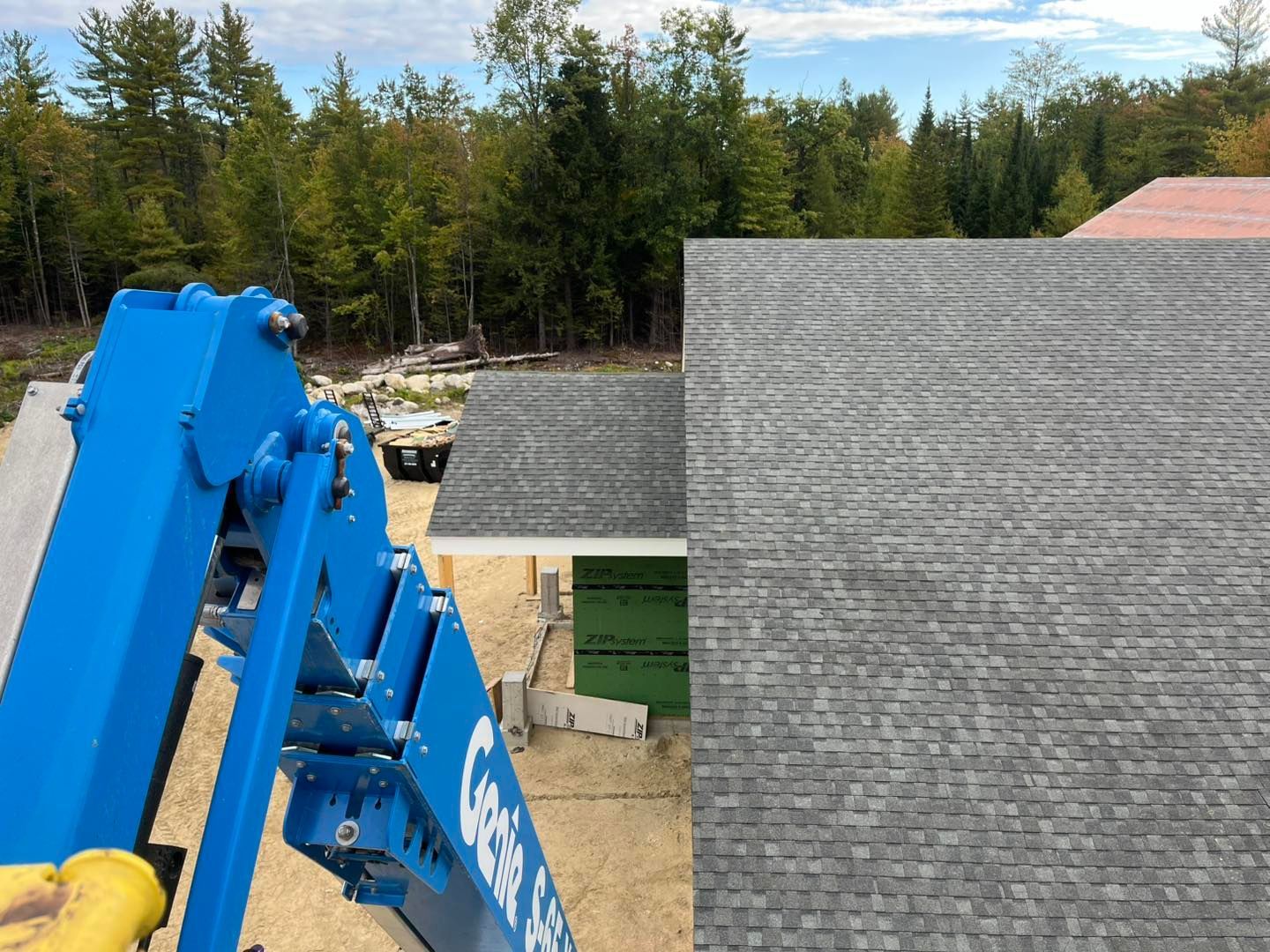 A view from a blue boom lift looking down at a house roof with gray shingles under construction, surrounded by woods.
