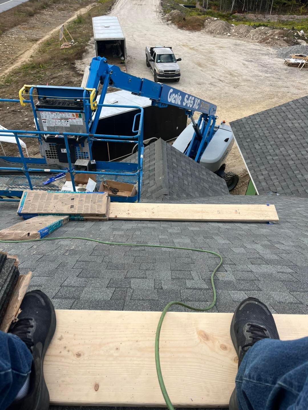A high-angle view from a roof showing a person's feet on a scaffold plank, with a blue boom lift parked below.