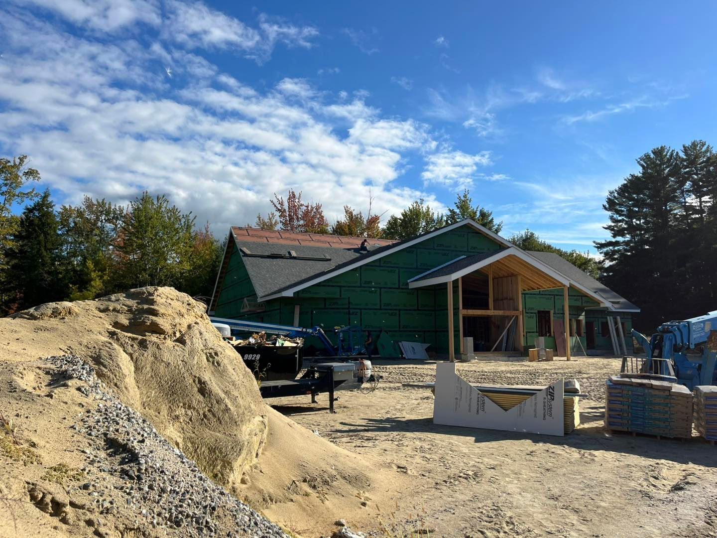 A residential house under construction, featuring green exterior sheathing, a partially shingled roof, and a sandy lot.