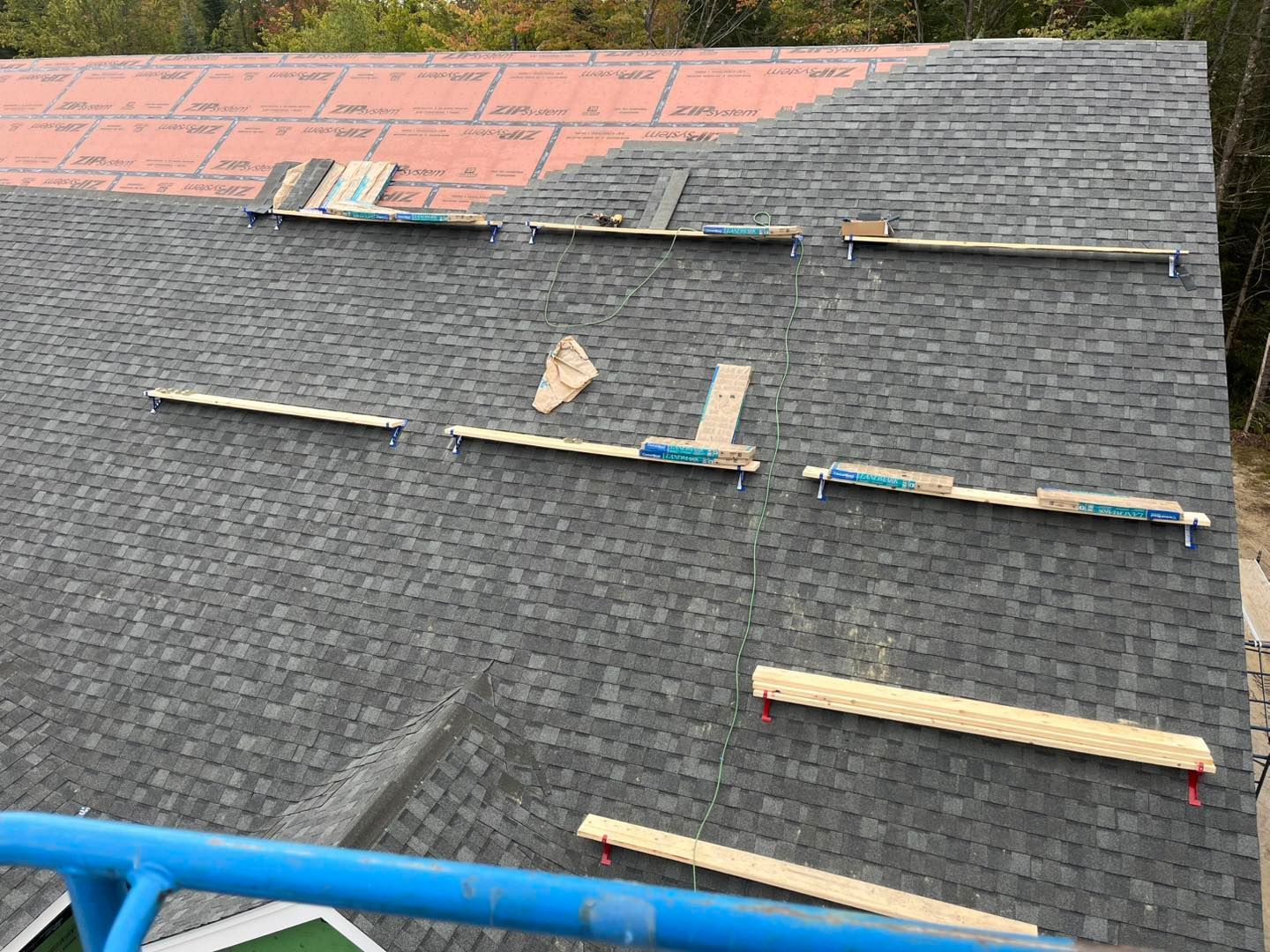 A steep residential roof under construction with grey shingles installed over red underlayment and wooden safety boards.