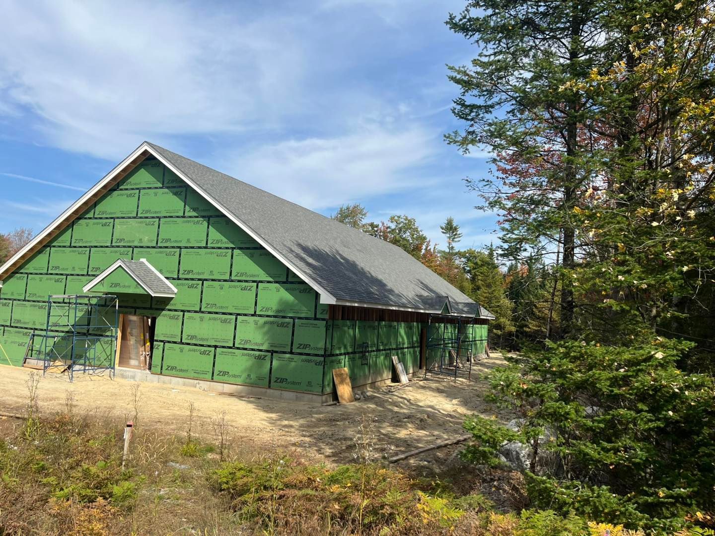 A partially constructed building covered in green exterior sheathing panels, nestled in a wooded area under a blue sky.