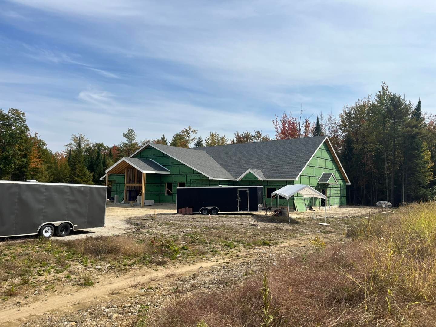 A newly constructed house with green sheathing and a dark shingled roof sits on a dirt lot near a forest under a blue sky.