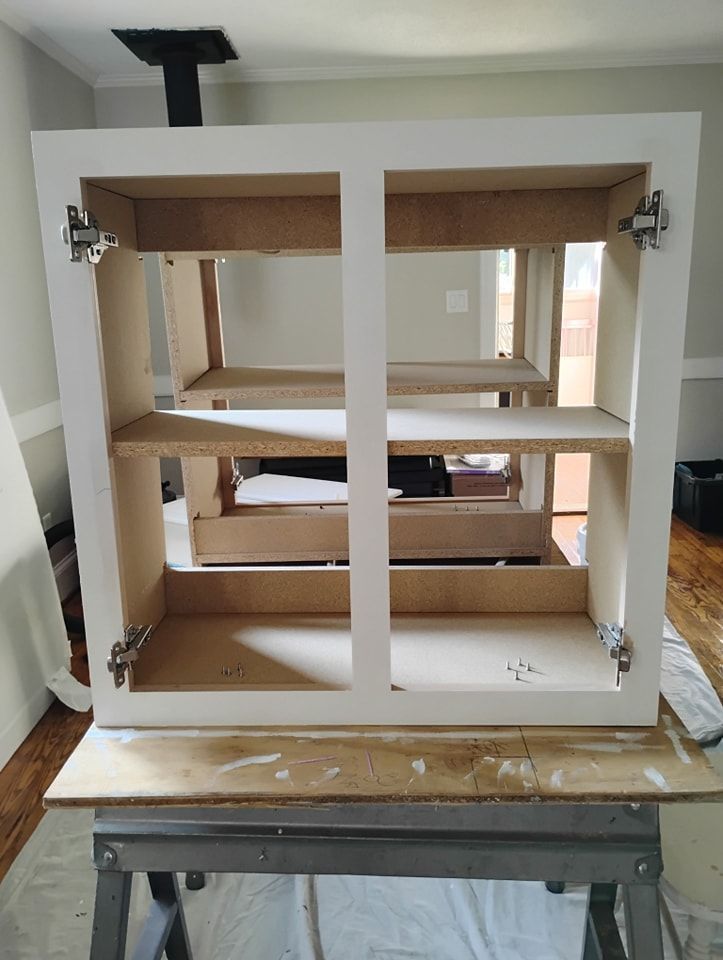 A white, unfinished wooden kitchen cabinet frame with metal hinges sits on a workbench in a room.