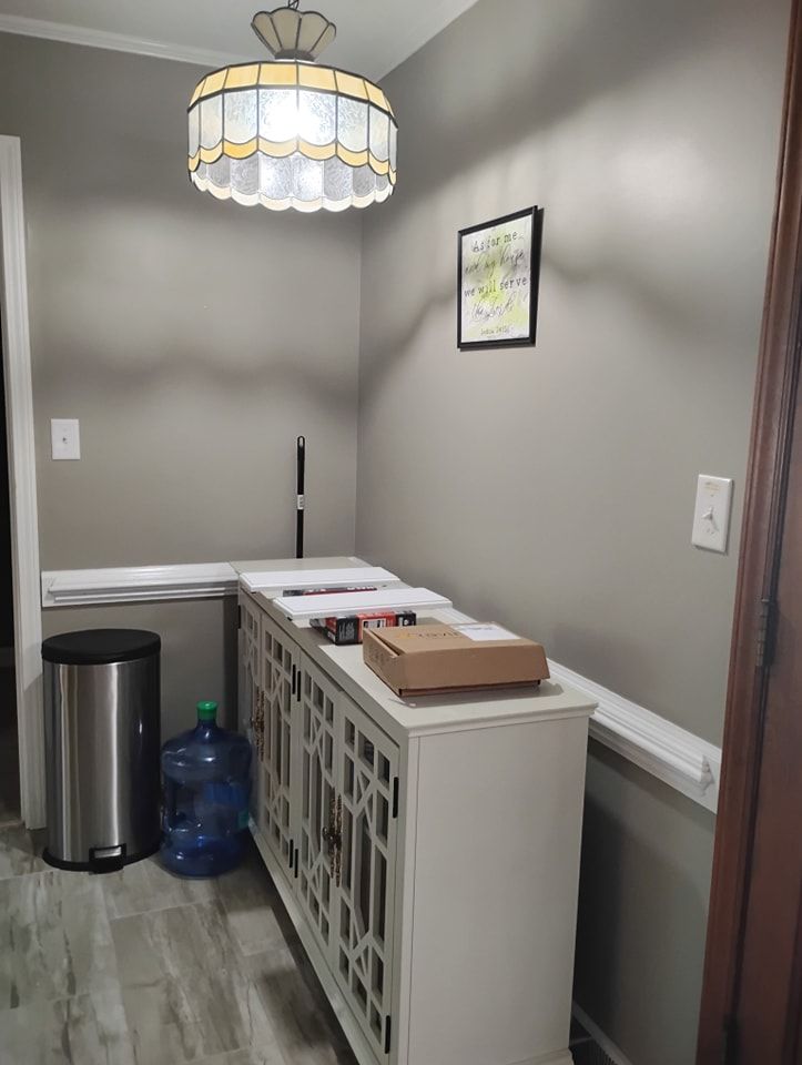 A small hallway with light gray walls, a white lattice-front cabinet, a stainless steel trash can, and a hanging lamp.