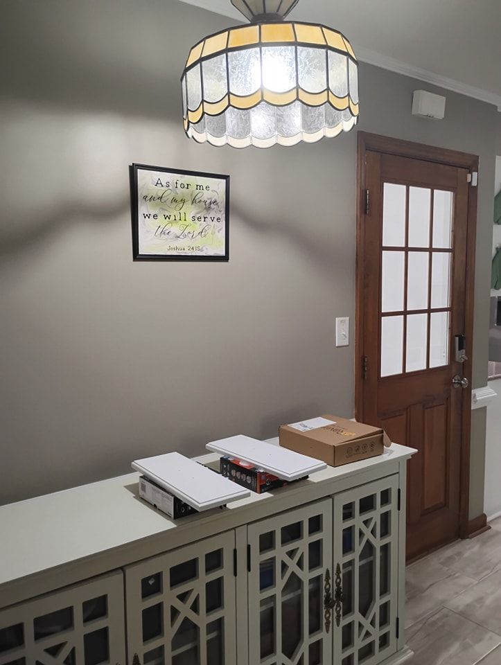 A light-colored cabinet stands against a gray wall near a wood-framed door, topped with packages under a pendant light.