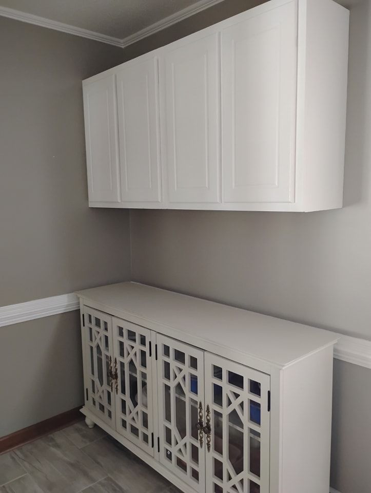 A white cabinet with glass-paned doors sits below white wall-mounted storage cabinets against a light gray wall.