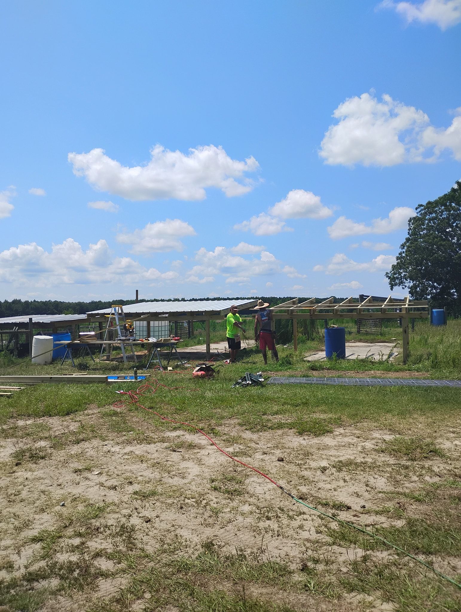 Two people work under a partially constructed wooden shelter in an open, sandy field under a blue sky with scattered clouds.