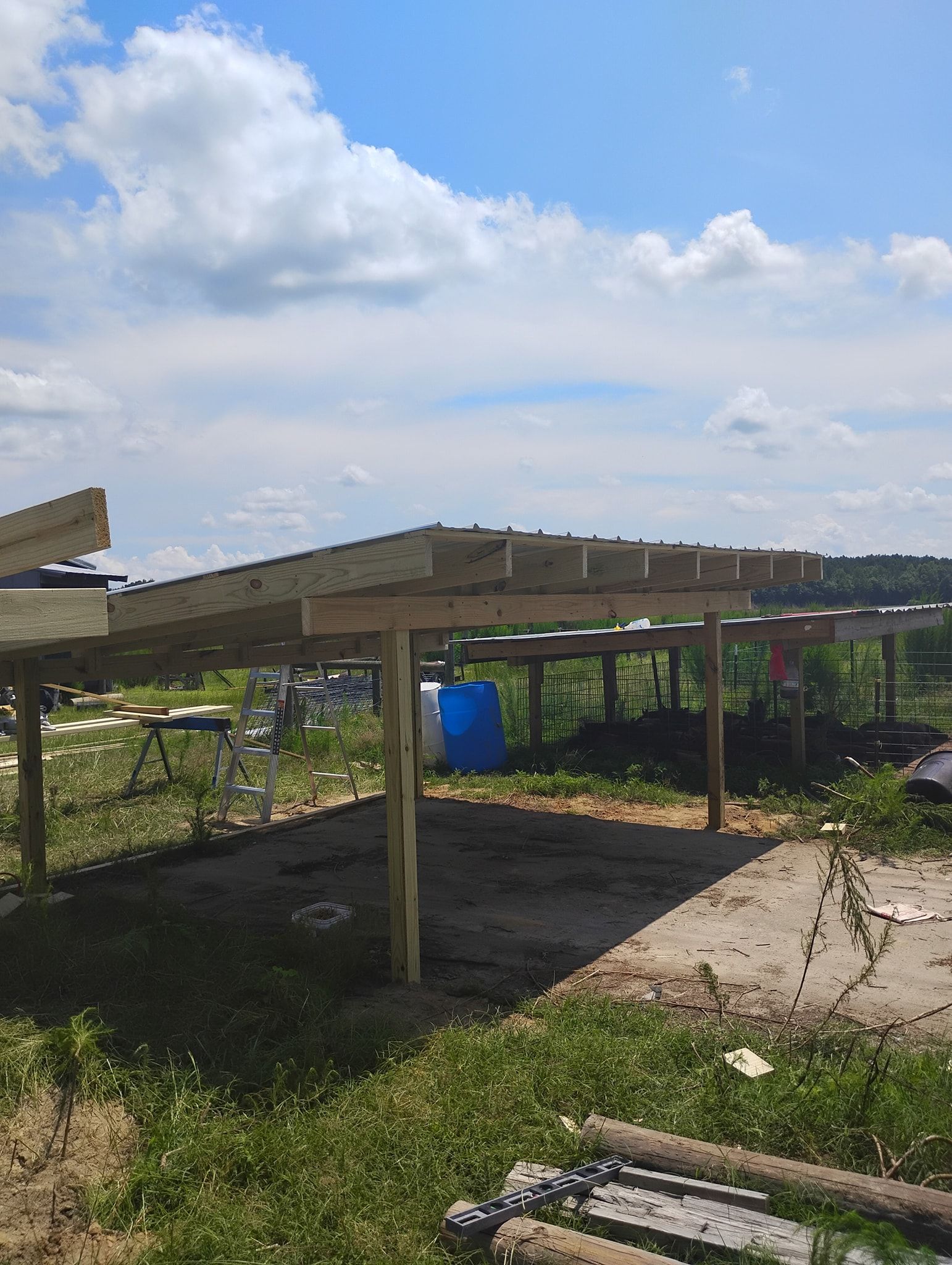 Wooden frame of an open-sided shelter under construction in a grassy field under a sunny blue sky.