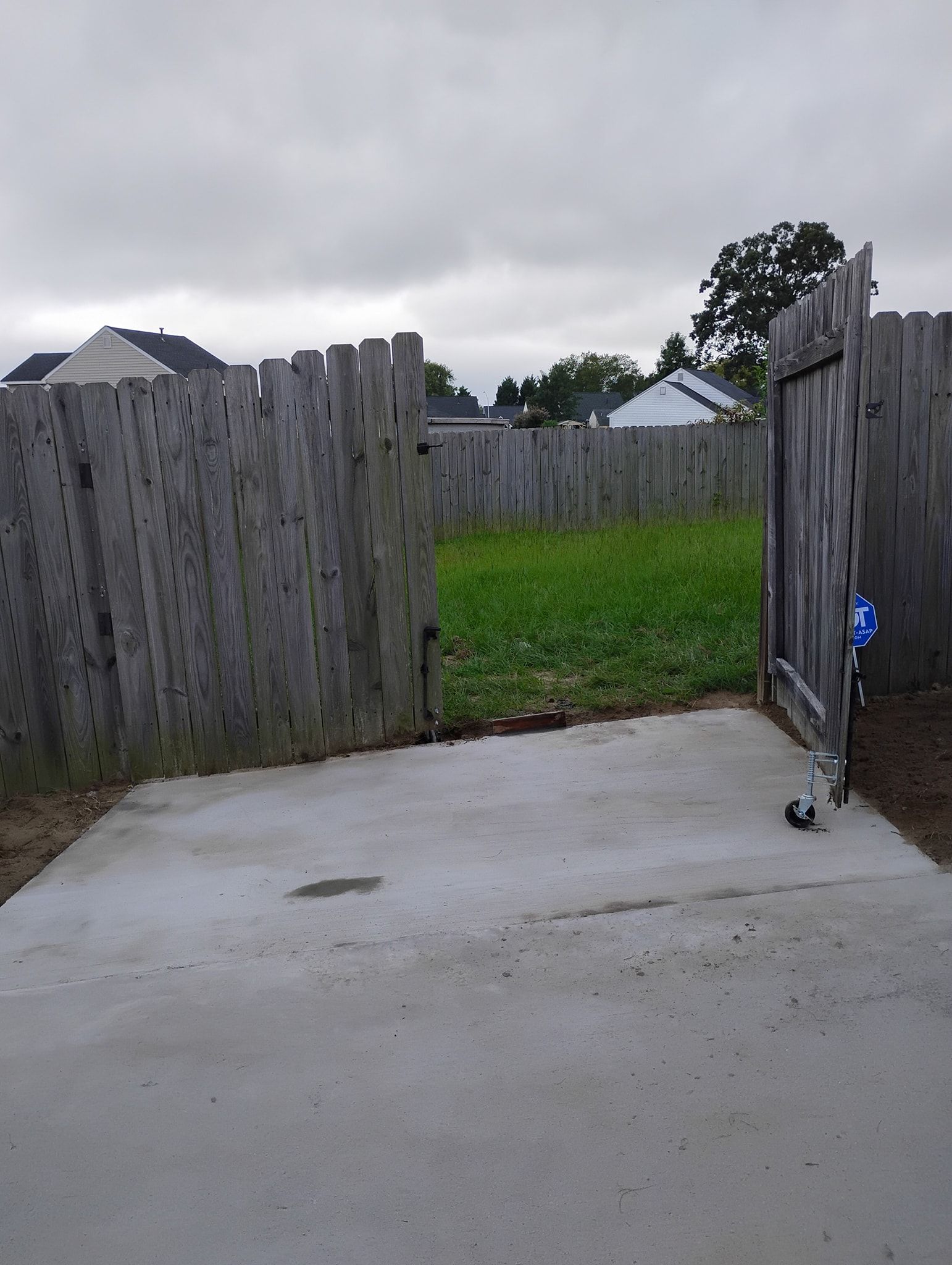 A concrete driveway leading to a gated gap in a weathered wooden fence, opening into a grassy backyard under a cloudy sky.