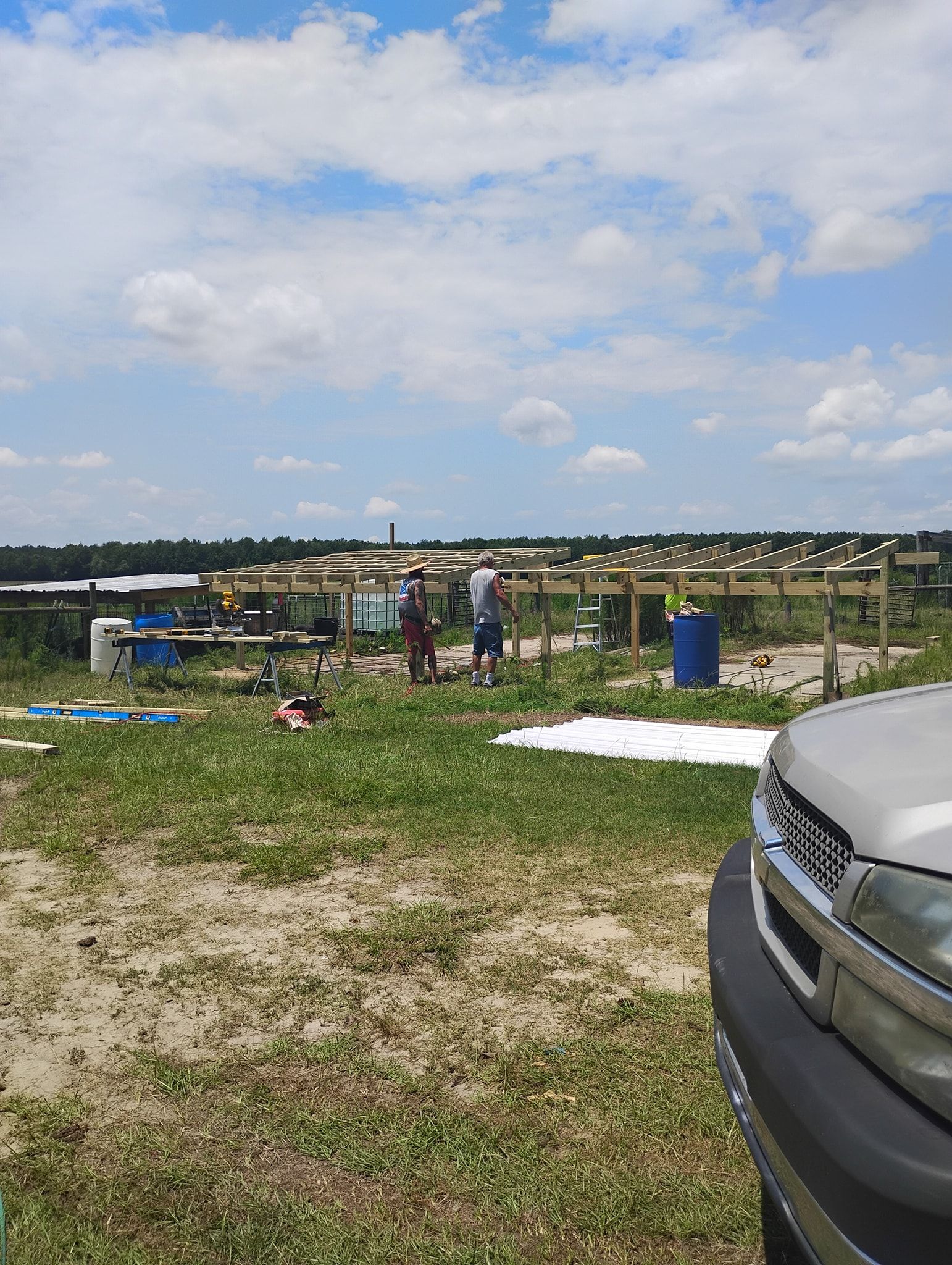 Two people stand near wooden framework structures in a sunny, grassy field, with a vehicle visible in the foreground.