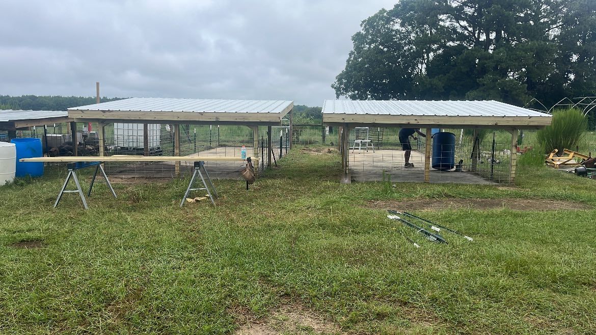 Two open-sided structures with metal roofs stand in a grassy field under a cloudy sky, with construction equipment nearby.