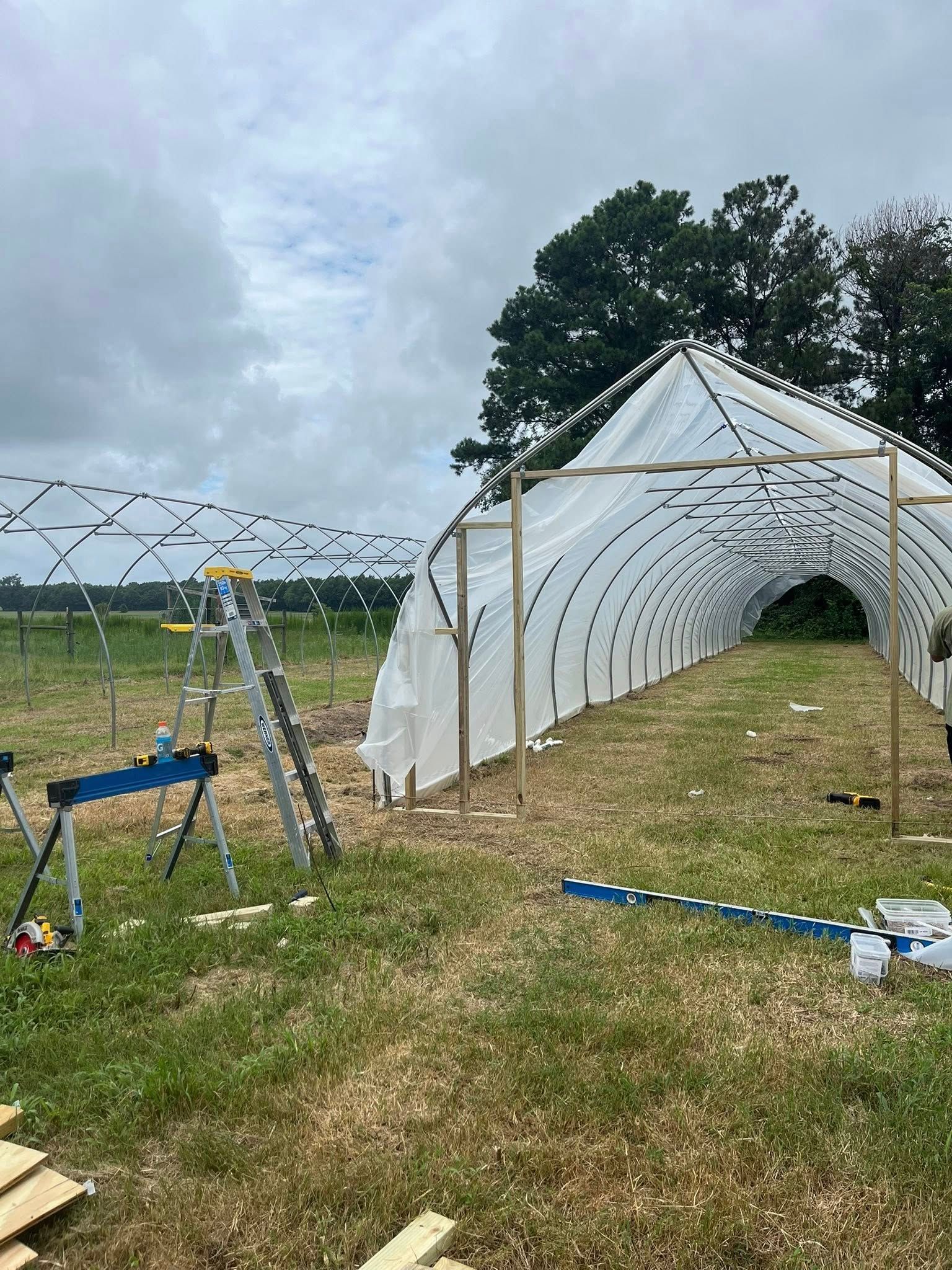 A partially constructed greenhouse frame with white plastic sheeting stands in a grassy field on a cloudy day.