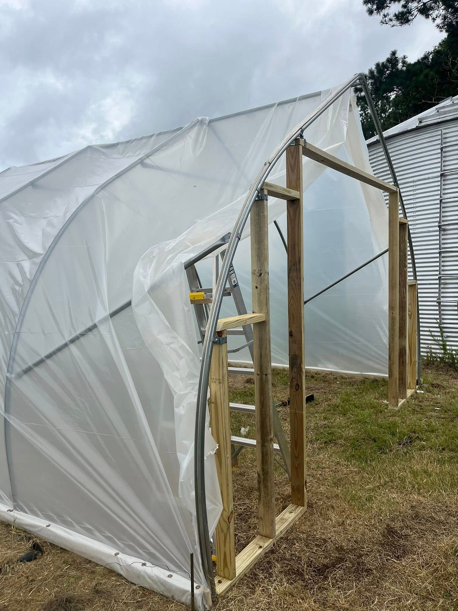 A half-constructed greenhouse with a wooden frame and clear plastic sheeting in a grassy yard.