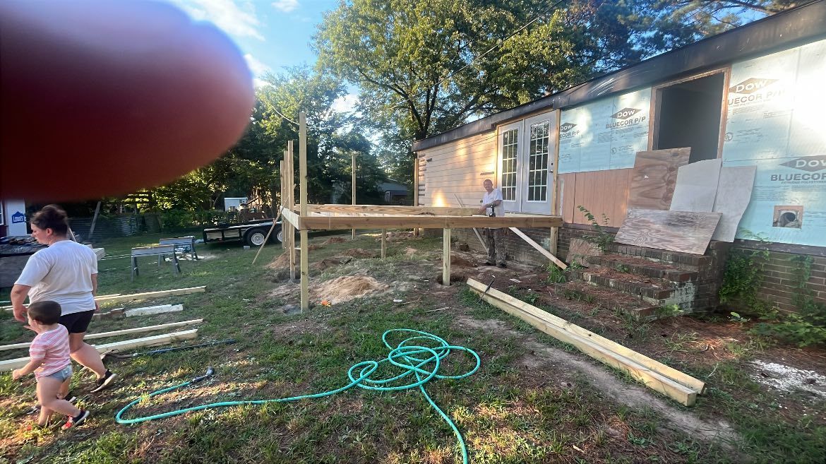 A person building a wooden deck attached to the exterior of a house, with a family walking in the yard nearby.