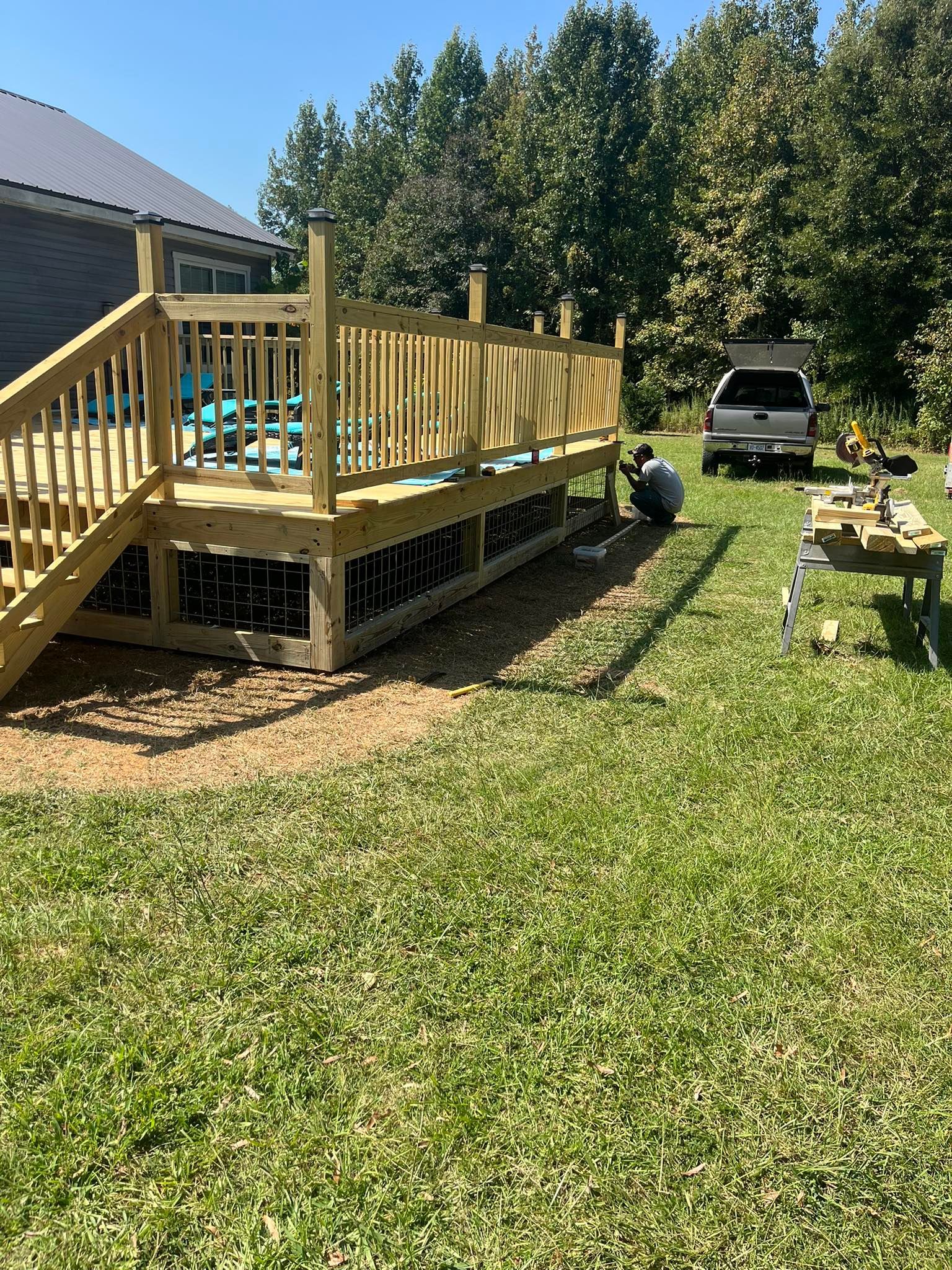 A person works on the base of a newly constructed wooden deck in a grassy backyard with a parked truck in the background.