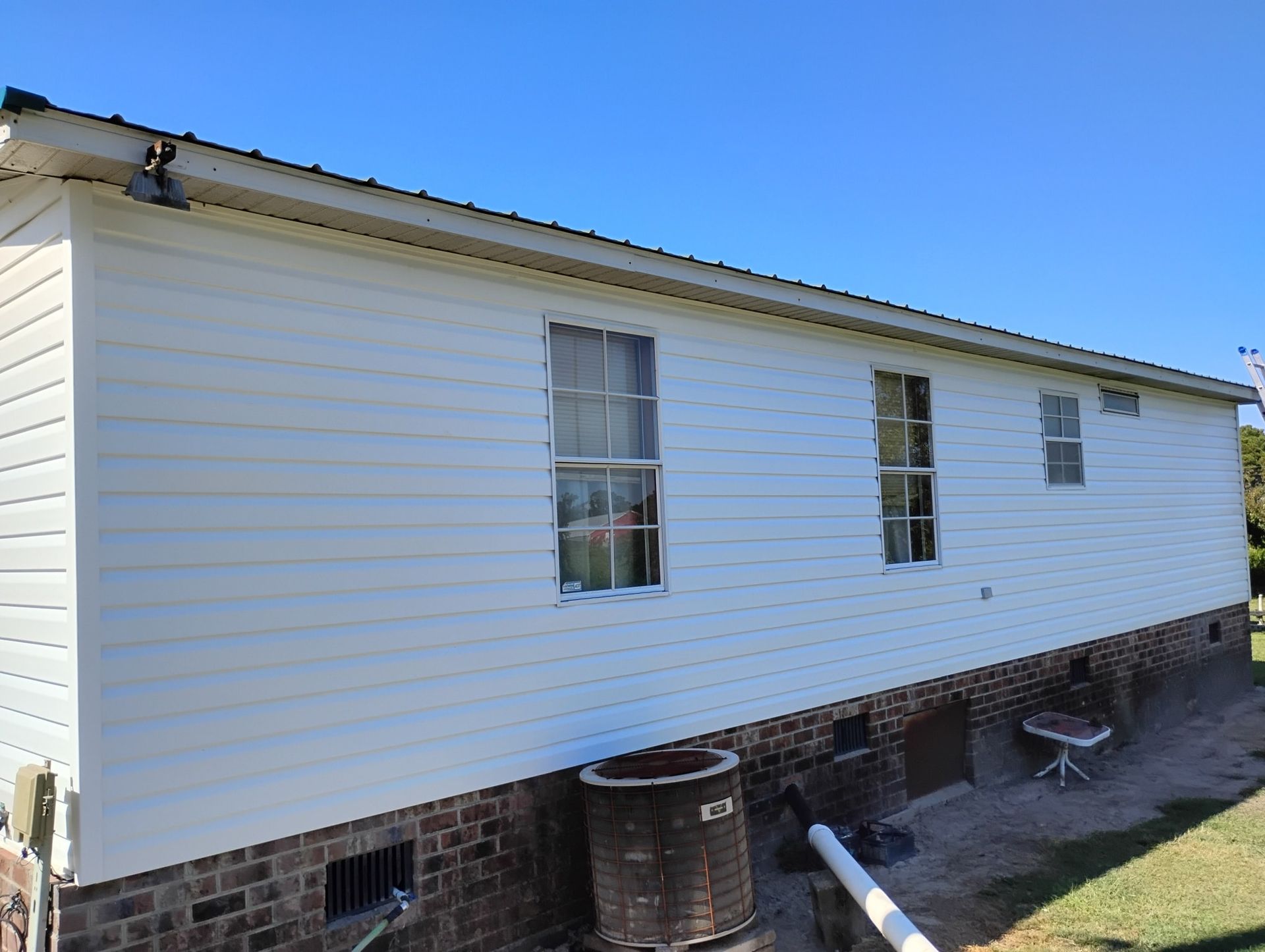 A white house with horizontal siding and three windows, featuring a brick foundation and a grassy yard under a blue sky.
