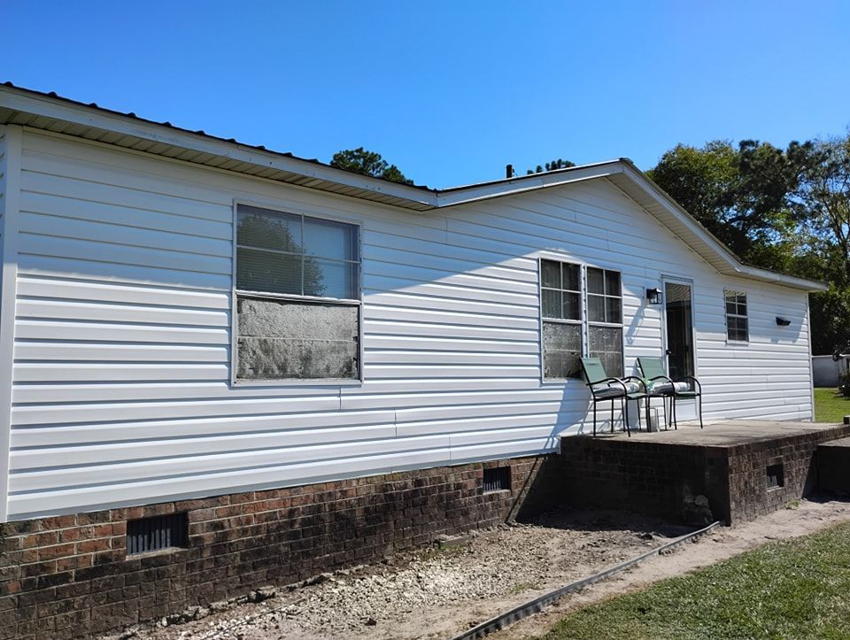 A white, single-story house with horizontal siding and a dark brick foundation, featuring a porch with two patio chairs.
