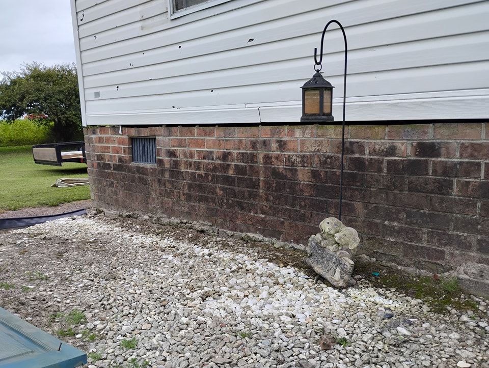 A brick foundation wall of a white-sided building with a metal lantern on a shepherd's hook and gravel ground.