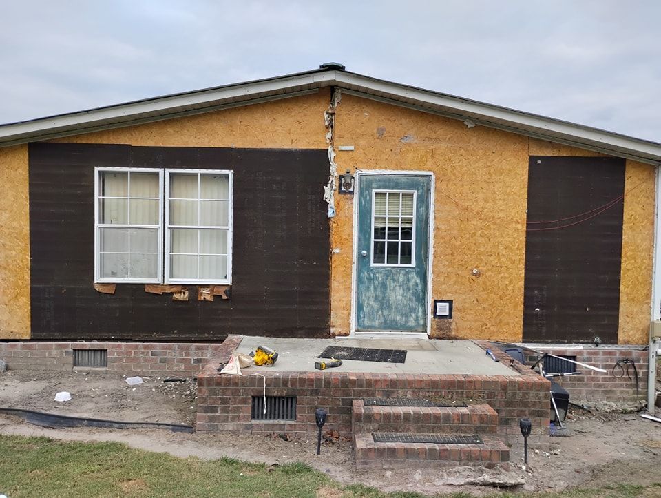 A single-story house exterior under renovation, featuring exposed plywood siding, a brick foundation, and a central door.